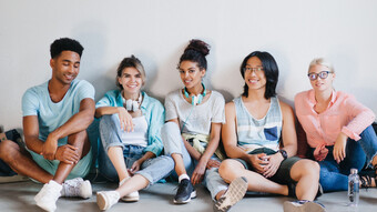 Heureux étudiants portant des baskets élégantes et des accessoires assis ensemble sur le sol avec les jambes croisées. Jeunes passionnés de nationalités différentes se détendant dans une pièce lumineuse et riant.